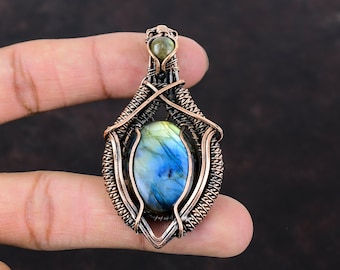 May include: A handcrafted pendant featuring a blue and green labradorite gemstone, encased in a copper wire wrap. The pendant has an oval shape with intricate wire detailing and a small, round stone at the top.