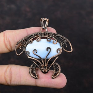Puede incluir: Un colgante envuelto en alambre de cobre con una piedra ovalada azul claro. El colgante tiene un diseño delicado e intrincado con espirales y bucles.