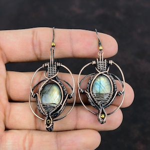 May include: A pair of copper wire wrapped earrings with a labradorite gemstone in each. The earrings are a dark copper color with black wire accents. The labradorite gemstones are iridescent and have a blue-green hue.