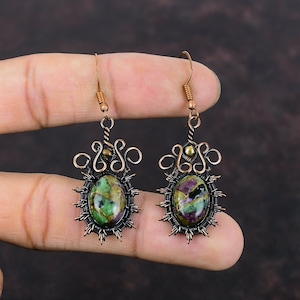 May include: A pair of copper wire wrapped earrings with green, purple, and black gemstone accents. The earrings feature a delicate, intricate design with a small gold bead at the top.