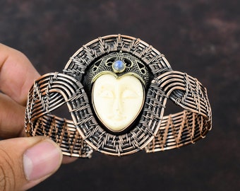 May include: A copper wire woven bracelet with a white face cameo and a blue gemstone in the center. The bracelet is made of two different colored wires, one copper and one silver.