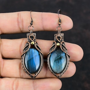 May include: A pair of copper wire wrapped earrings with blue labradorite gemstones. The earrings have a teardrop shape and are accented with gold beads.