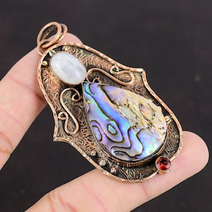 May include: A copper pendant featuring a pearlescent abalone shell and a white moonstone. The pendant is intricately designed with floral patterns and has a red garnet accent.