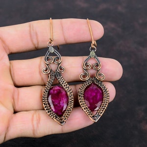 May include: A pair of dangle earrings with a pink gemstone set in a copper wire frame. The earrings have a delicate, intricate design with a swirl at the top.