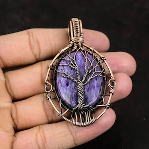 Pode incluir: Um pingente de fio de cobre envolto em torno de uma pedra de ágata dendrítica roxa e azul em forma de árvore. O pingente tem formato oval e possui uma argola de cobre.
