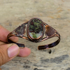 May include: A copper bracelet with a green and brown oval stone set in the center. The bracelet has two heart-shaped cutouts on either side of the stone.