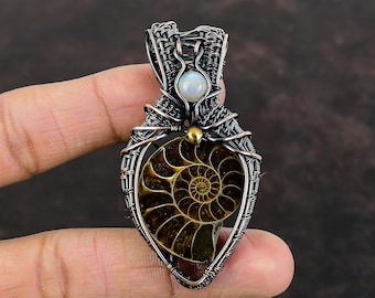Puede incluir: Un colgante de alambre envuelto en plata y cobre con un fósil de amonita y una piedra blanca iridiscente. El colgante tiene forma de corazón y tiene un acento dorado.