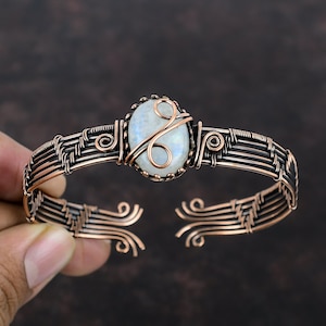 May include: A copper wire wrapped bracelet with a white moonstone cabochon. The bracelet is made with multiple strands of copper wire that are woven together to create a delicate and intricate design. The moonstone is set in a bezel setting and is surrounded by a copper wire frame.