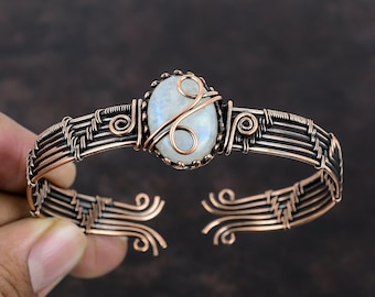 May include: A copper wire wrapped bracelet with a white moonstone cabochon. The bracelet is made with multiple strands of copper wire that are woven together to create a delicate and intricate design. The moonstone is set in a bezel setting and is surrounded by a copper wire frame.