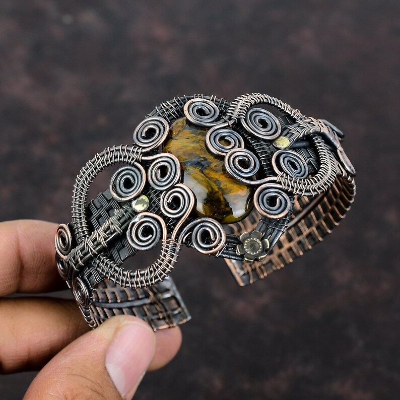 May include: A copper wire wrapped bracelet with a large tiger's eye gemstone in the center. The bracelet is made of multiple wire coils and has a woven texture.