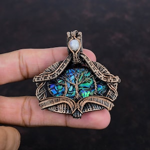 Op de afbeelding: Handgemaakte koperdraad hanger met een boomontwerp, iriserende abalone schelp en een maansteen accent. De hanger is ingewikkeld geweven en meet ongeveer 5 cm in hoogte en 4 cm in breedte.