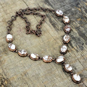 May include: A copper-toned necklace featuring oval white and brown stones. The stones are set in a copper-colored metal and connected by a chain. The necklace has a Y-shaped design with a pendant of three stones.