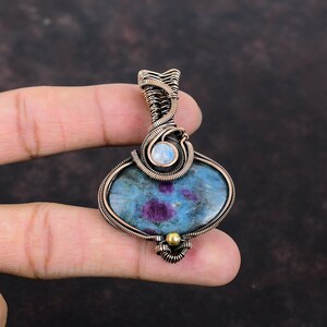 May include: A copper wire wrapped pendant featuring a blue and purple gemstone with a smaller white moonstone accent. The pendant has a gold bead at the bottom.