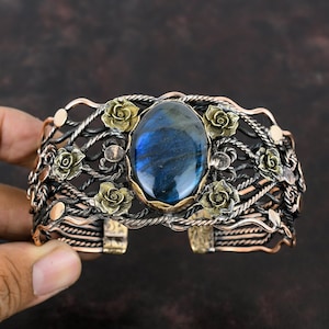 Puede incluir: Brazalete de puño ornamentado con una gran piedra ovalada de labradorita azul. El brazalete presenta un diseño calado detallado con rosas de color bronce y detalles plateados y cobrizos. Una joya única.