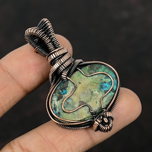 May include: A copper wire wrapped pendant featuring a green and blue gemstone with a unique pattern. The pendant is a handmade piece of jewelry.