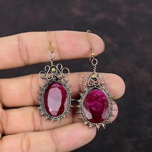 以下が含まれることがあります： 大きな楕円形の深い赤色の宝石をあしらった、手作りのイヤリングのペア。 宝石は、複雑なワイヤーワークと装飾的なアクセントを備えた銅色の金属フレームにセットされています。 イヤリングはフック留めです。