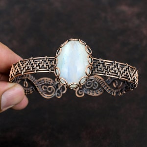 May include: A handcrafted copper bracelet featuring a large, oval-shaped moonstone. The moonstone is light blue and surrounded by a decorative copper setting. The bracelet has a woven design and intricate scrollwork.