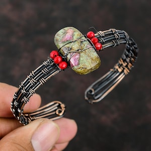 May include: A copper wire bracelet featuring a green and pink stone and red beads. The bracelet is woven in a criss-cross pattern.