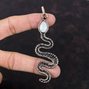 以下が含まれることがあります： 白い虹色の宝石が付いた銅線製の蛇のペンダント。蛇は巻き付いており、繊細で複雑なデザインです。