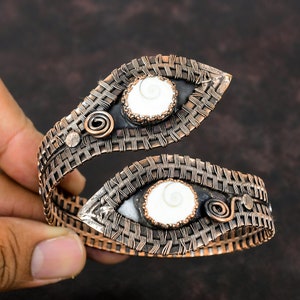 May include: A copper wire wrapped bracelet with two white shell inlays. The bracelet is woven in a basket weave pattern and has a slightly curved shape.