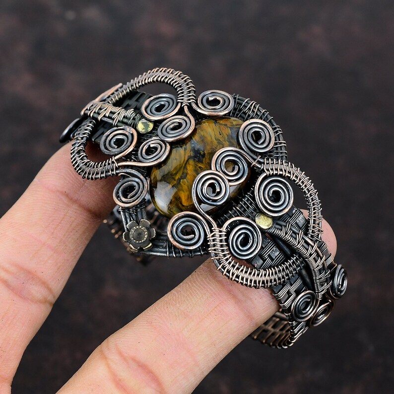May include: A copper wire wrapped bracelet with a large tiger's eye gemstone in the center. The bracelet is made of multiple coils of wire and has a dark oxidized finish.
