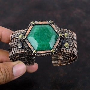 May include: A copper wire cuff bracelet with a large green gemstone in the center. The gemstone is set in a hexagonal frame with small gold accents.