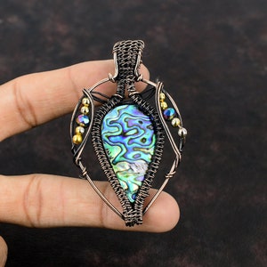 Op de afbeelding: Een hanger van koperdraad met een traanvormige abalone-schelp met iriserende blauwe, groene en paarse tinten. De hanger is versierd met gouden en zwarte kralen.