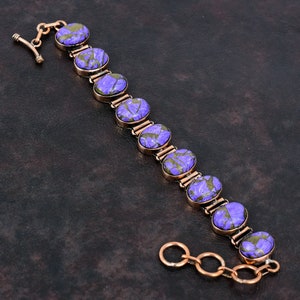 Puede incluir: Un brazalete de color cobre con piedras ovaladas moradas y verdes. Las piedras están engastadas en un metal color cobre y conectadas por pequeños eslabones de cobre. El brazalete tiene un cierre de palanca.