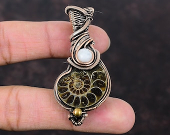 Puede incluir: Un colgante envuelto en alambre de cobre con un amonite fosilizado y un acento de perla blanca. El colgante es una pieza de joyería única e intrincada.