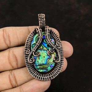 Op de afbeelding: Een hanger omwikkeld met koperdraad met een iriserende abalone-schelp. De hanger is ovaalvormig en heeft een gedetailleerd draadontwerp.