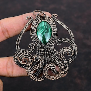 Könnte beinhalten: Handgefertigter Kupferdraht-Anhänger mit einem ovalen Malachitstein. Der Anhänger hat ein aufwendiges Design mit wirbelnden Drahtdetails und einer dunklen Patina. Der Malachitstein ist ein leuchtendes Grün mit einzigartigen Mustern.