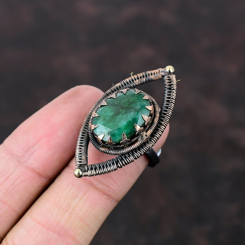 May include: A green gemstone ring with a copper and black wire design. The ring is shaped like an eye.
