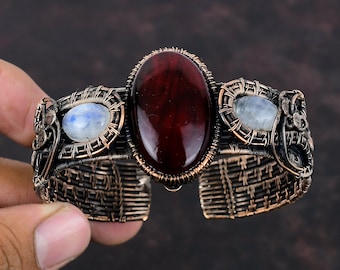 May include: A copper wire wrapped bracelet with a large red gemstone and two smaller moonstone gemstones.