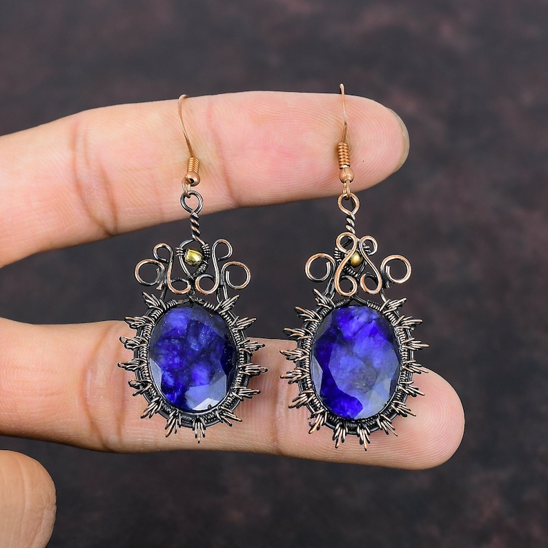 May include: A pair of handcrafted earrings featuring large, oval-shaped, deep blue gemstones. The gemstones are set in a dark metal frame with a sunburst design. The earrings have copper-colored wire accents and ear hooks.