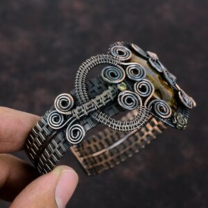 May include: A silver and copper wire wrapped bracelet with a brown and gold tiger's eye stone. The bracelet is made with a woven wire design and features several wire coils.