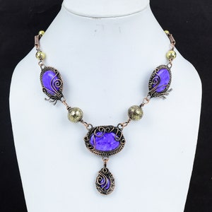May include: A handcrafted necklace featuring a series of oval and teardrop-shaped purple stones. The stones are set in ornate, dark copper-colored wirework. The necklace includes brass-colored beads and a clasp.
