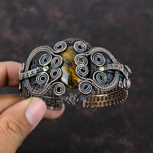 May include: A copper wire wrapped bracelet with a brown and gold tiger's eye gemstone. The bracelet is decorated with intricate wire coils and has a woven texture.