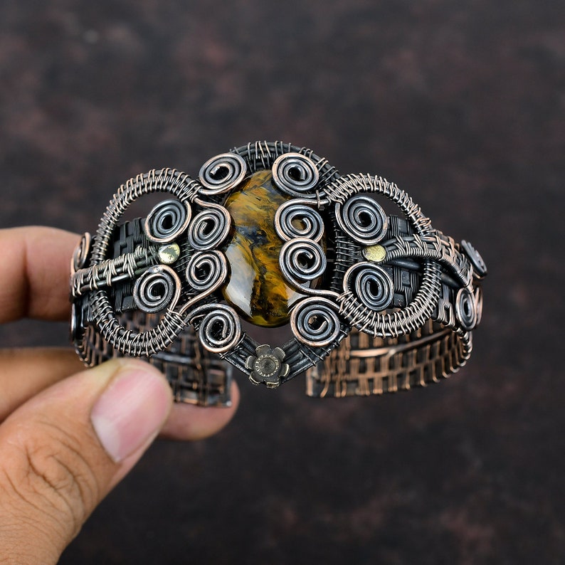 May include: A copper wire wrapped bracelet with a brown and gold tiger's eye gemstone. The bracelet is decorated with intricate wire coils and has a woven texture.