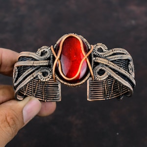 May include: A copper wire wrapped bracelet with a red gemstone cabochon in the center. The bracelet is made of copper wire and has a delicate, intricate design.
