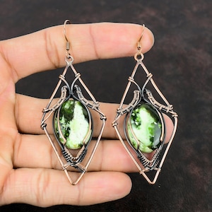 Peut inclure: Une paire de boucles d'oreilles en pierre verte et blanche avec du fil de cuivre et noir enroulé autour des pierres. Les boucles d'oreilles ont la forme d'un diamant et ont un design suspendu.