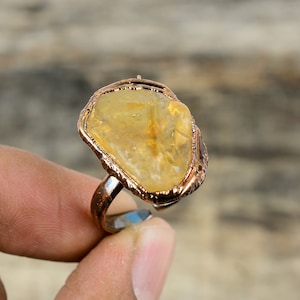 May include: A copper wire wrapped ring with a large, yellow citrine gemstone. The ring is on a finger.