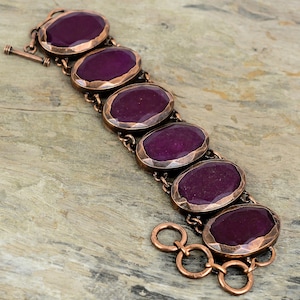 May include: A copper bracelet with seven oval-shaped purple stones. The stones are set in copper bezels and the bracelet has a lobster clasp closure.