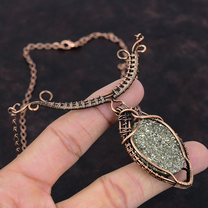 K&ouml;nnte beinhalten: Handgefertigte Kupferdraht-Halskette mit einem gro&szlig;en, oval geformten Pyritstein. Die Halskette hat eine zarte Kette und ein einzigartiges, k&uuml;nstlerisches Design. Der Pyritstein hat ein metallisches, gesprenkeltes Aussehen.