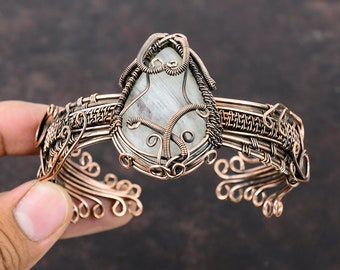 May include: A copper wire wrapped bracelet with a white gemstone in the center. The bracelet is made of intricate wirework and has a delicate, feminine design.
