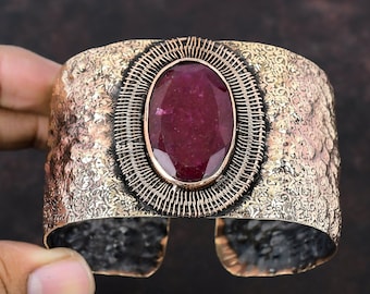May include: A wide copper cuff bracelet featuring a large oval-shaped red gemstone set in a wire cage design. The bracelet has a hammered texture and a floral pattern.