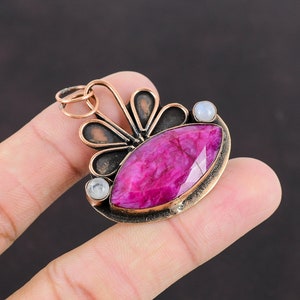 May include: A handcrafted pendant featuring a large, faceted, magenta-colored gemstone as the focal point. The pendant has a copper-colored metal setting with a decorative, openwork design and two small, round, white gemstones. The pendant is approximately 1.5 inches long.