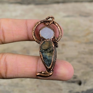 Puede incluir: Un colgante de alambre de cobre envuelto con una piedra preciosa azul claro y verde y una piedra preciosa blanca en el centro.