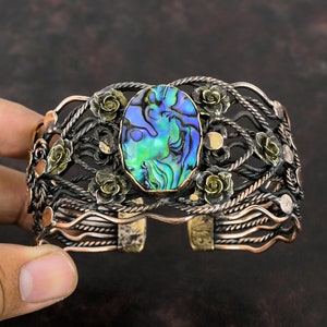 Op de afbeelding: Een handgemaakte manchetarmband met een ovaal abalone schelpmiddenstuk met iriserende blauwe en groene tinten. Het metaalwerk omvat ingewikkelde bloemen- en touwachtige ontwerpen in zilver, koper en goud, waardoor een uniek, artistiek accessoire ontstaat.