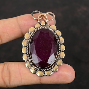 Puede incluir: Un colgante grande de forma ovalada con una piedra preciosa de color púrpura intenso engastada en un marco de metal dorado. El marco tiene un diseño decorativo con pequeños acentos redondos.