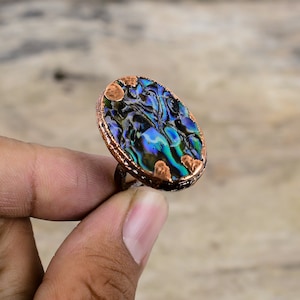 Pode incluir: Um anel de cobre com uma incrustação oval de concha de abalone. A concha tem cores iridescentes azuis, verdes e roxas.